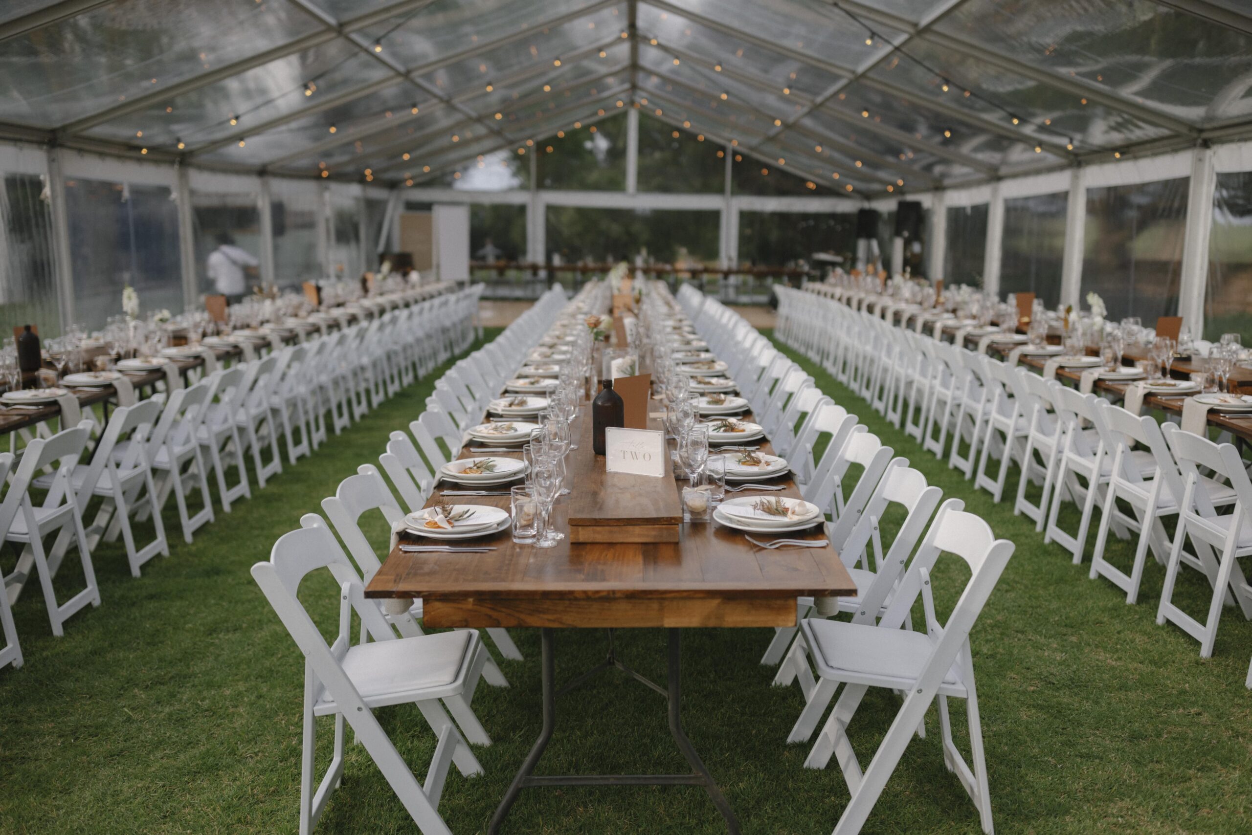 Reception - clear marquee with long rows of wooden tables with white chairs, fairy lights and minimalistic table decor
