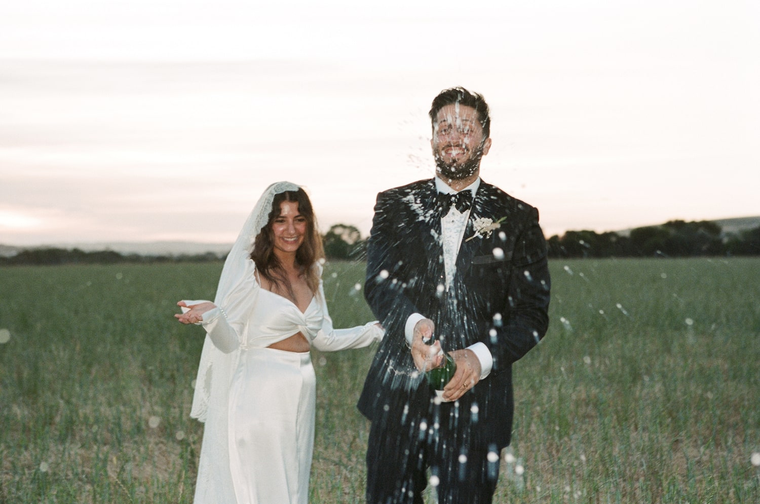 Bride and groom in a field during dusk, popping a bottle of bubbly