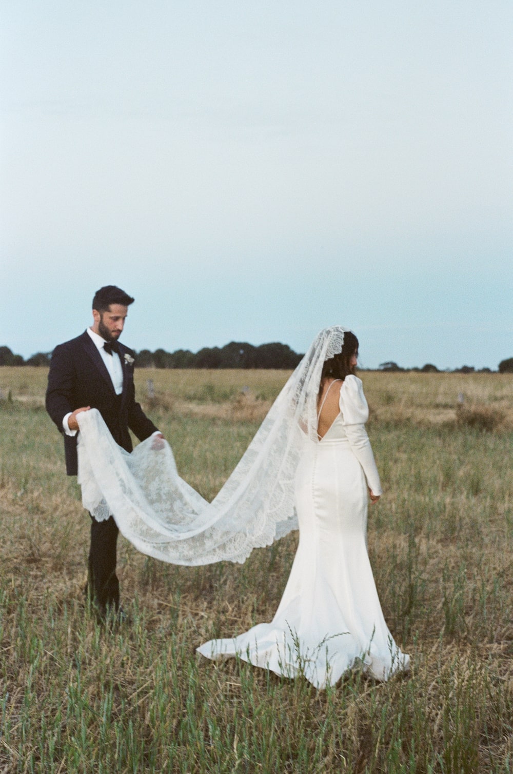 Bride and groom portrait in a field. Groom is holding brides mantilla veil up off the ground.