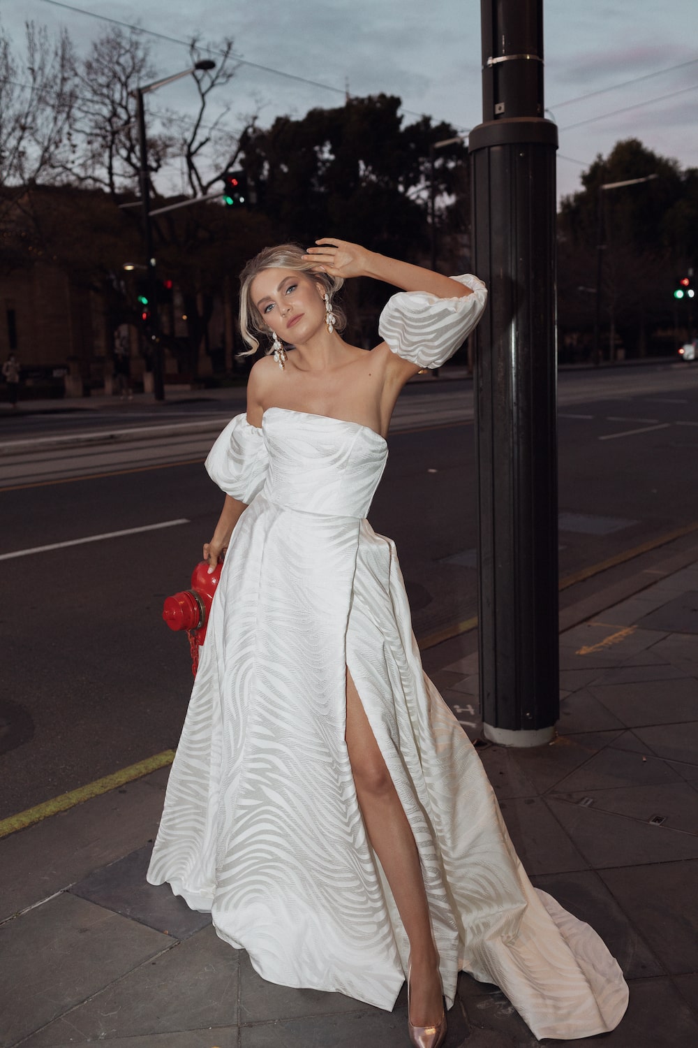 Model on sidewalk next to fire hydrant at night, posing with one arm up to her head, wearing the Concerto gown