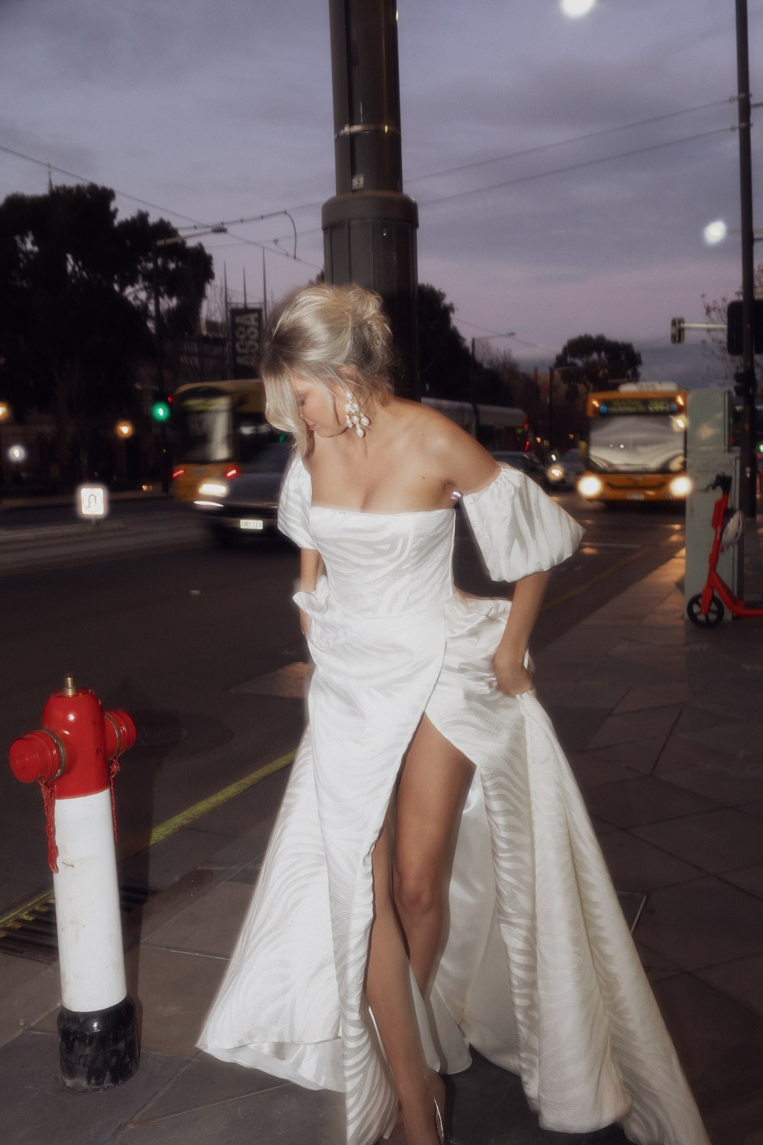 Model on sidewalk next to a fire hydrant at night, hitching skirt of the Concerto gown up to show off the high leg split.