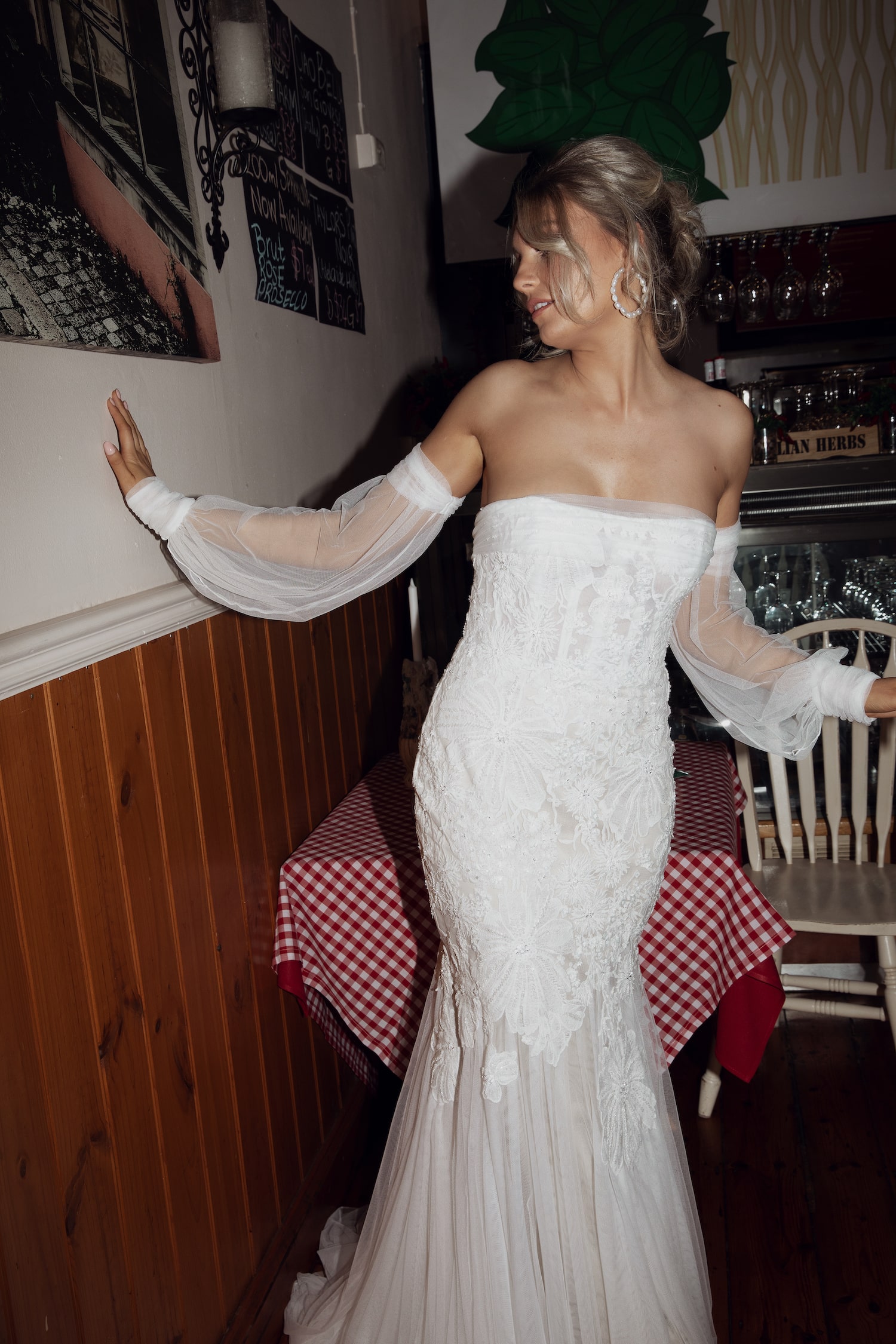 Model inside Italian cafe standing next to a small table with red and white check tablecloth leaning one hand against wall. Wearing the Verona gown.