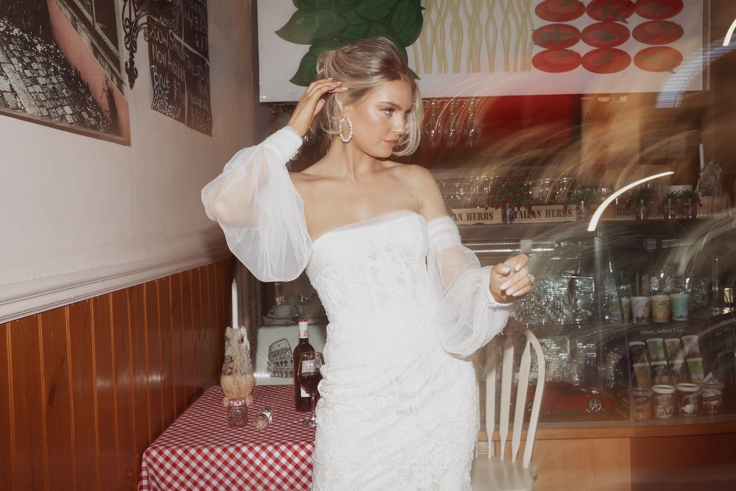 Model inside Italian cafe standing next to a small table with red and white check tablecloth. Wearing the Verona gown.
