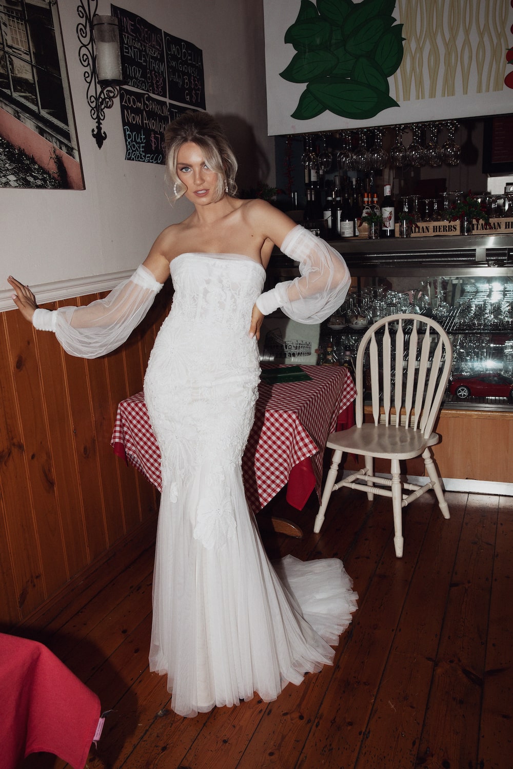 Model inside Italian cafe standing next to a small table with red and white check tablecloth leaning one hand against wall and one on waist. Wearing the Verona gown.