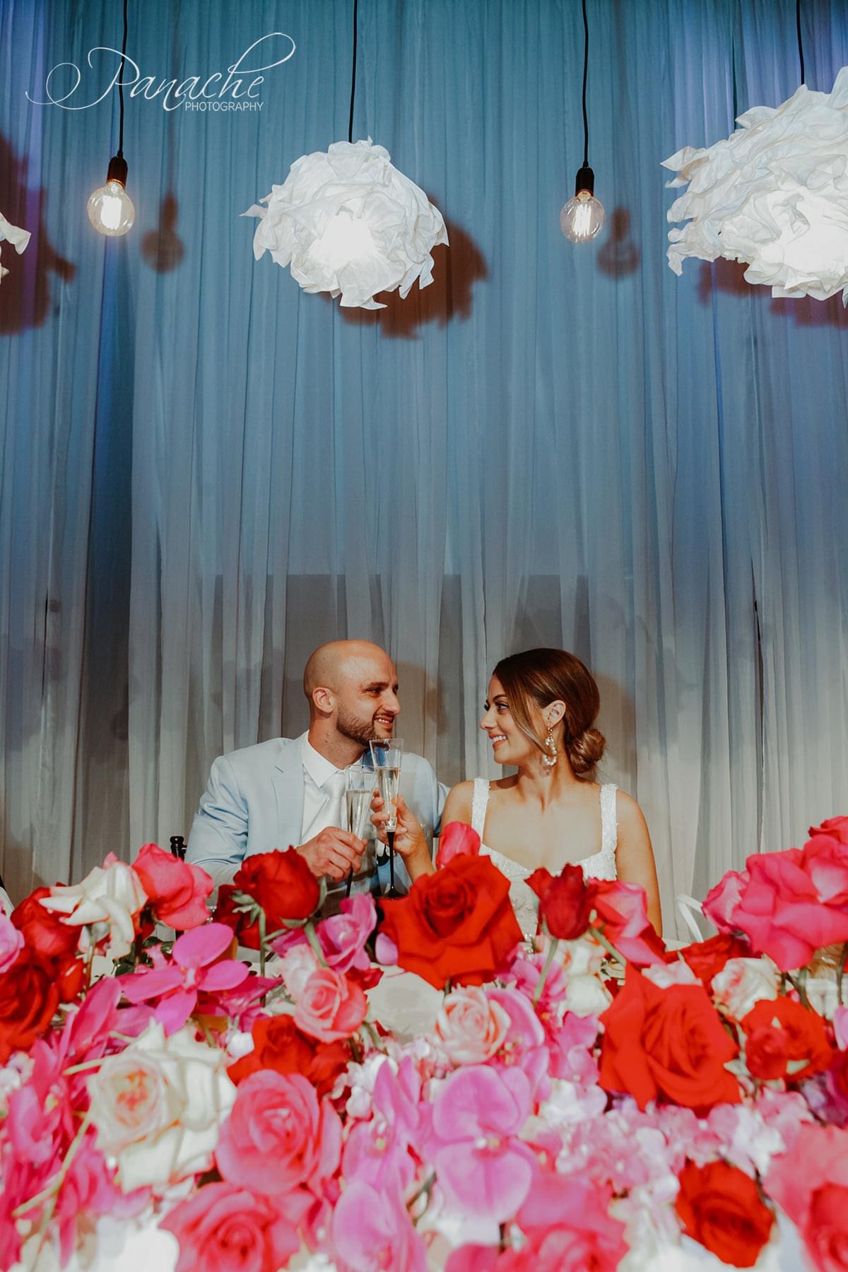 Cristiana and Jake sitting at their reception table. In front of the camera is lots of pink white and red florals.