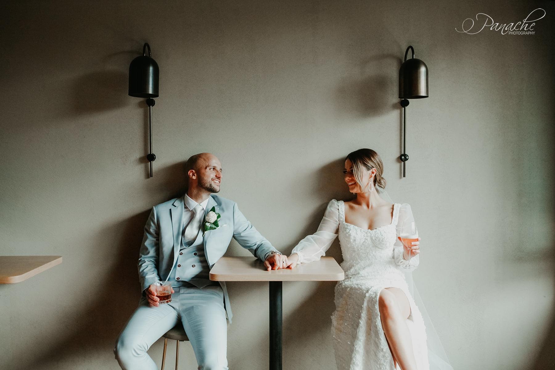 Newlyweds Cristiana and Jake sitting in a modern bar with a concrete wall backdrop, the couple are sitting at a high table with their backs against the wall holding hands across the table. Cristiana is also holding a cocktail in her other hand.