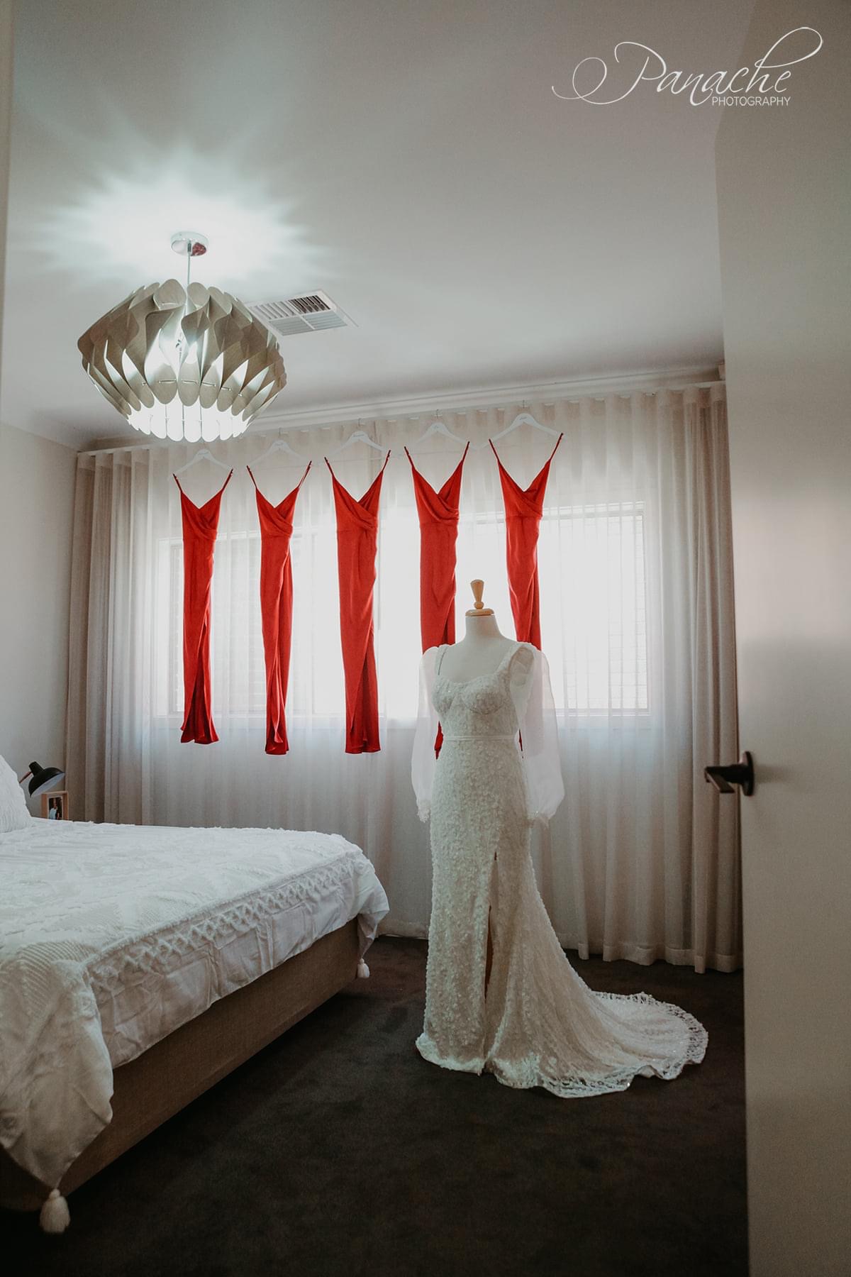 Cristiana's gown on a mannequin with red bridesmaids dresses hanging on a curtain rod in the background. Cristiana's gown is a slim mermaid gown in 3D flower embroidered tulle with an exposed corset cup bodice and detachable organza statement sleeves. 
