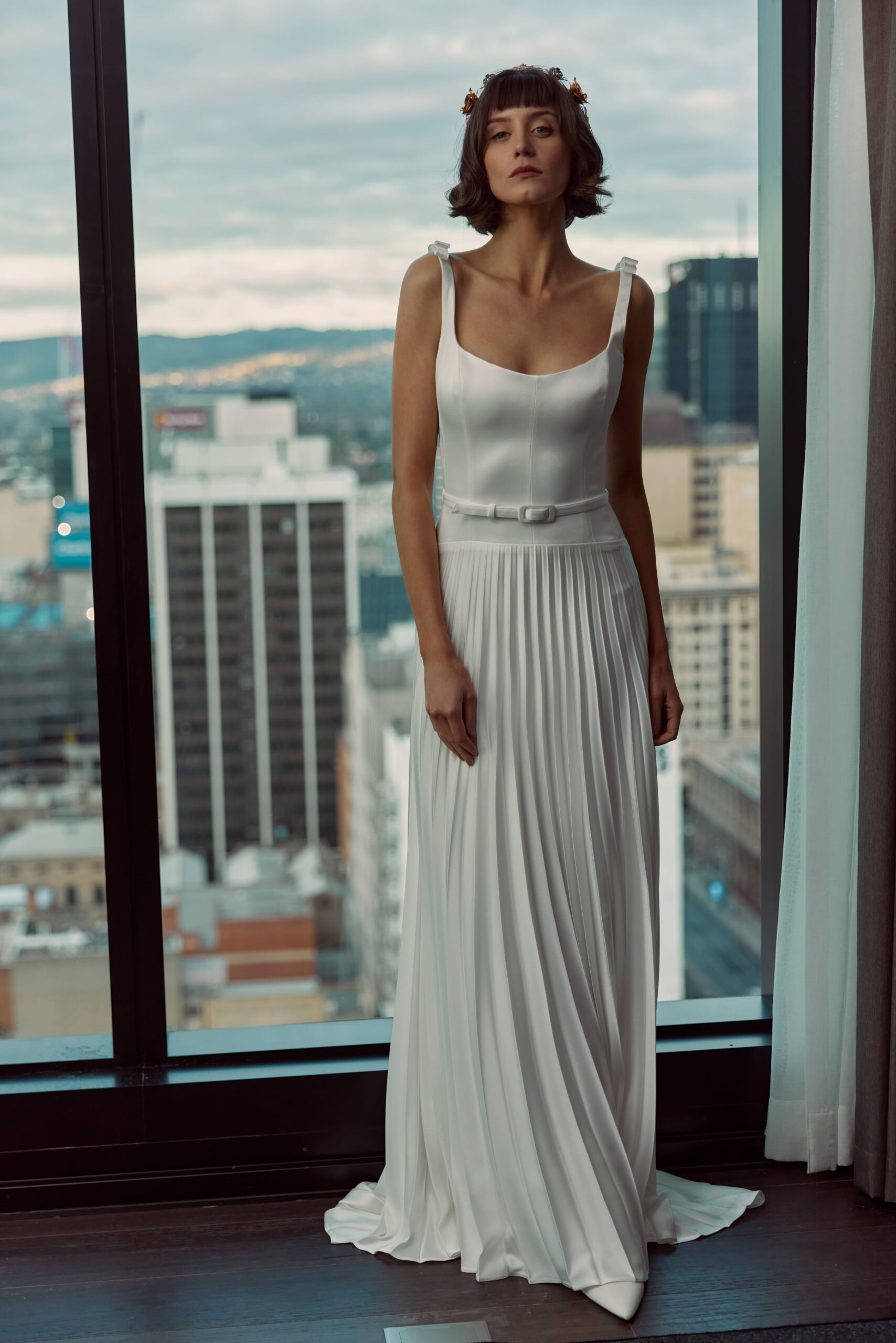 Lilou - An A-line gown featuring a drop waist, scoop neckline with straps, pleated skirt and a belt at the waist.