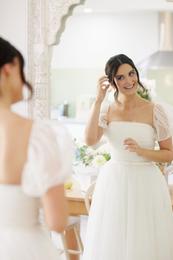 Maddy getting ready, wearing her gown and standing infront of a full length mirror putting in earrings