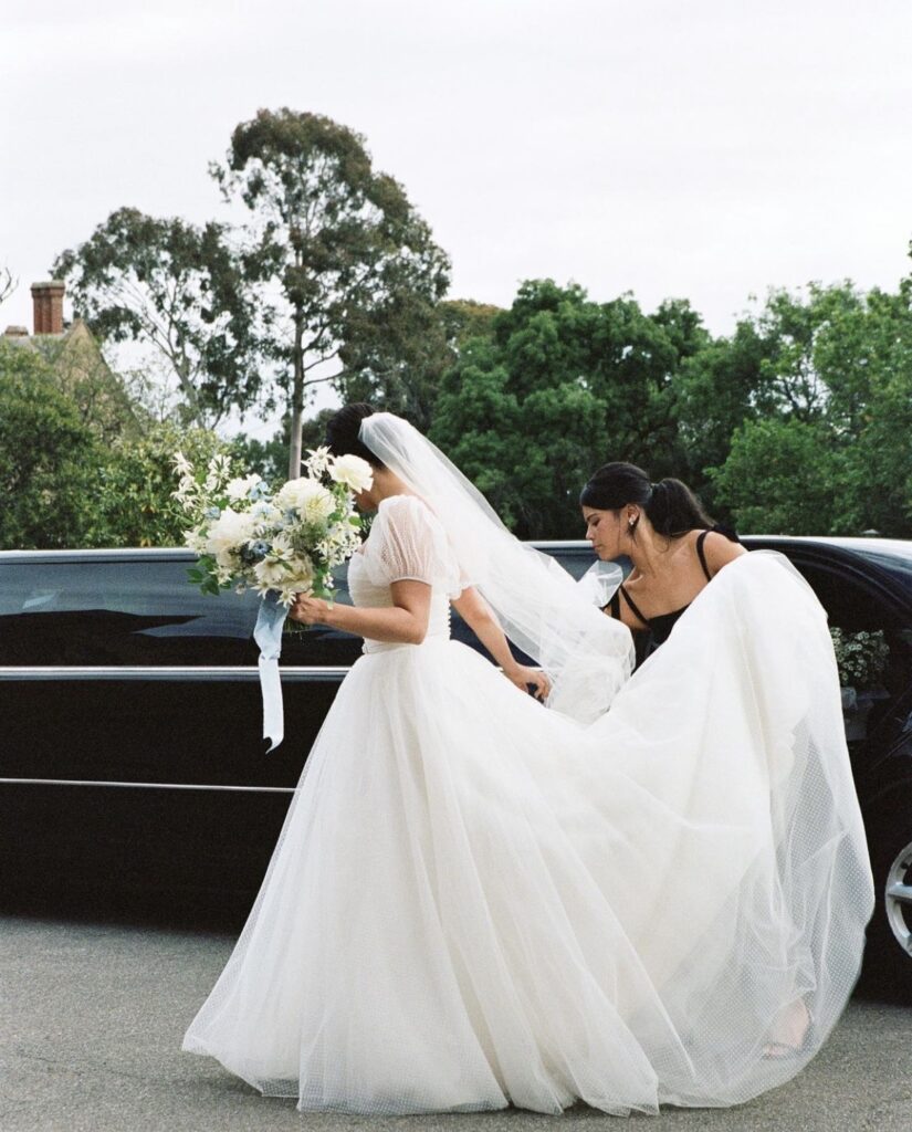 Maddy's bridesmaids holding up the train of her gown as she gets out of a limo holding her bouquet