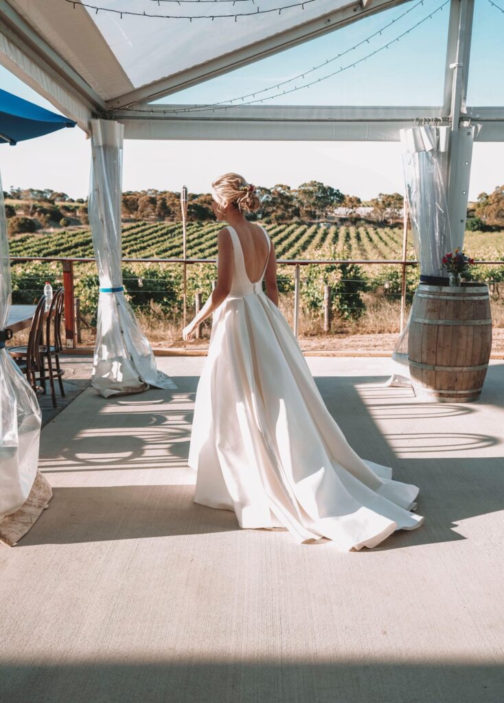 View of Maia in her gown from the back. A low square neckline. Views of vineyards behind.