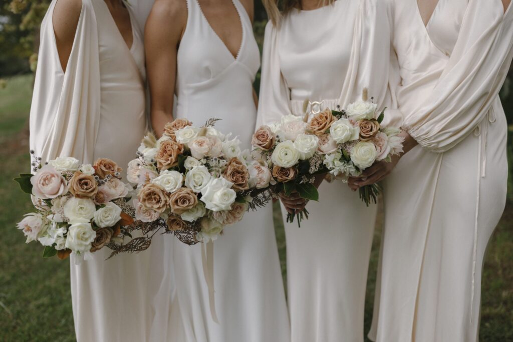 Close up of Olivia and bridesmaids gowns and bouquets - bridesmaids are wearing softly draped light creamy blush gowns with long sleeves. Bouquets are soft pink, cream and an earthy brown