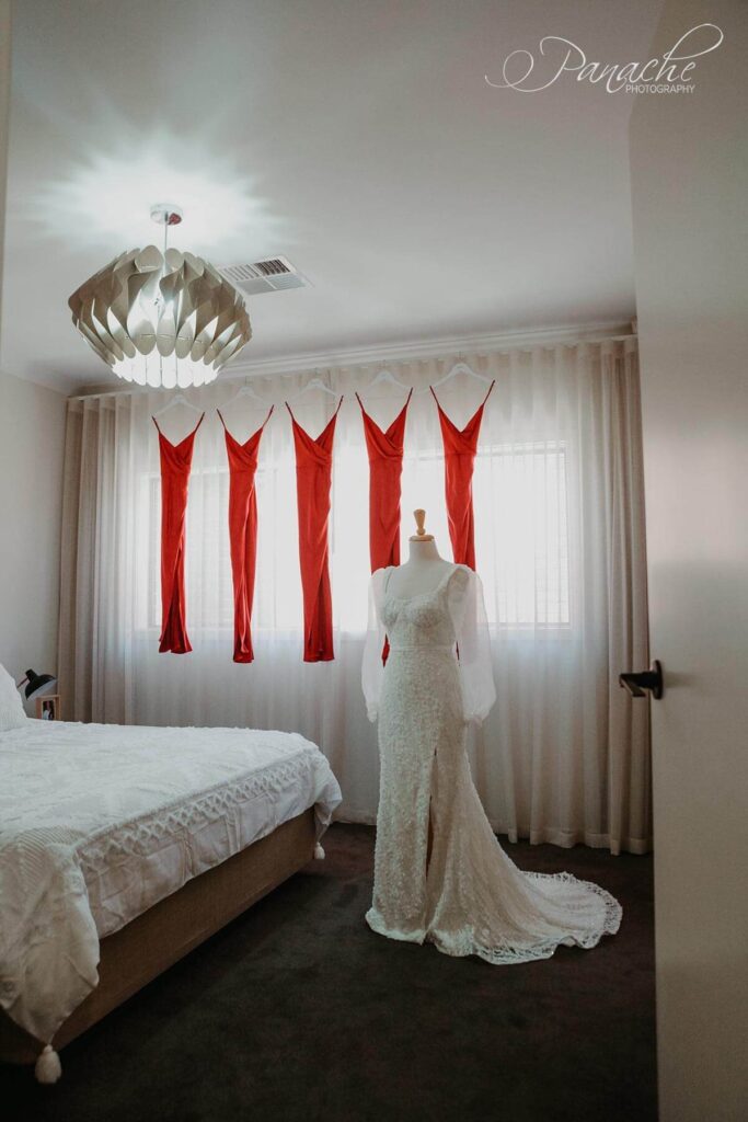 Cristiana's gown on a mannequin with red bridesmaids dresses hanging on a curtain rod in the background. Cristiana's gown is a slim mermaid gown in 3D flower embroidered tulle with an exposed corset cup bodice and detachable organza statement sleeves.