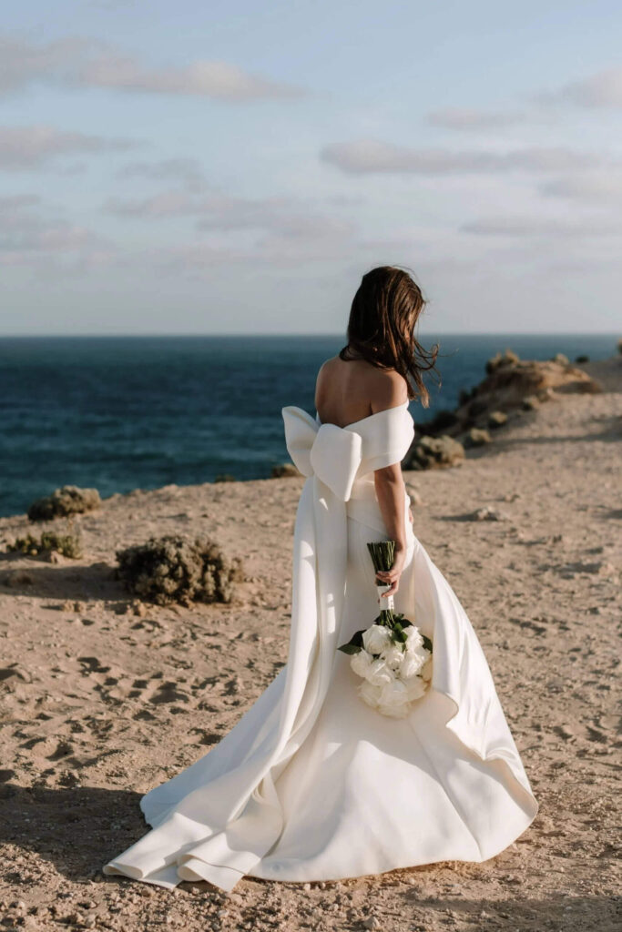 Georgia walking along a beach cliff, with bouquet in hand. Georgia's gown is figure-hugging, off the shoulder and has a big feature bow at the back.