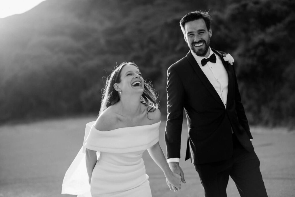 Real bride Georgia walking hand in hand with husband, laughing. Georgia's gown is figure-hugging, off the shoulder and has a big feature bow at the back.