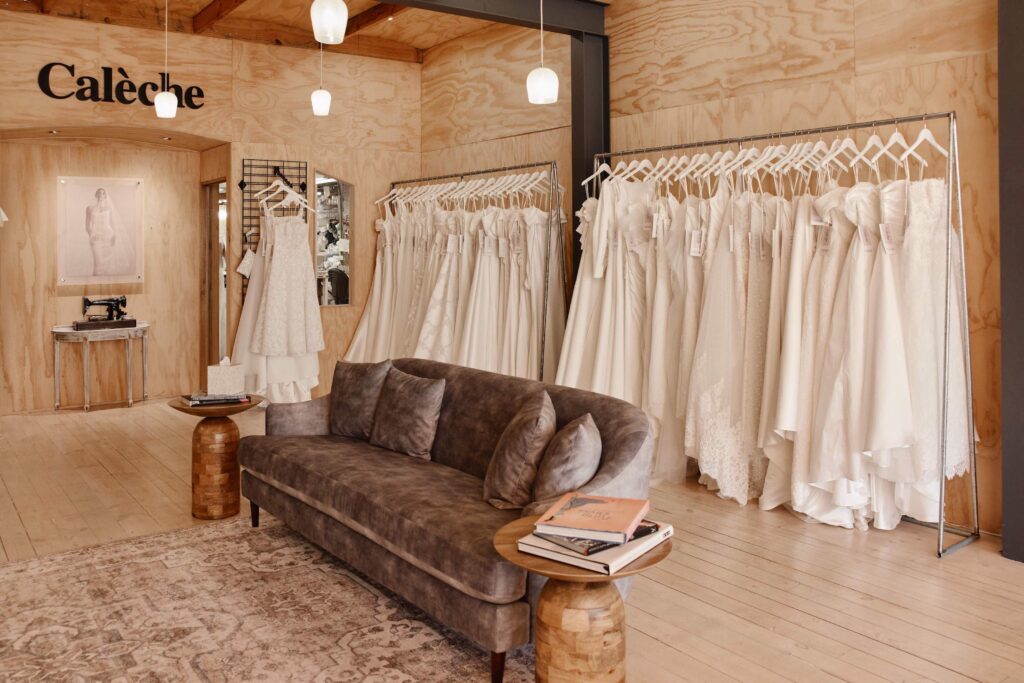 Wedding dress boutique showroom with two racks of bridal gowns, a grey couch and wooden side tables and patterned rug on the floor.